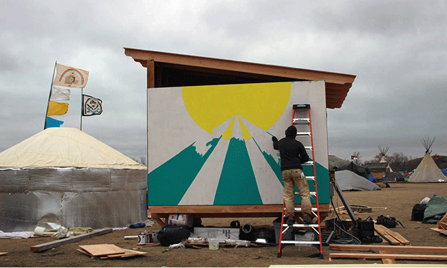 A volunteer paints the side of one of the cabins reassembled at the Standing Rock encampment from southwestern Oregon. Photo by Roger Peet.